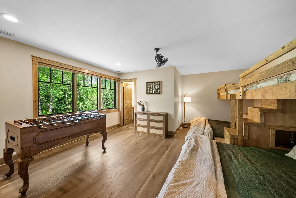 A spacious, playful bunk room featuring custom twin-over-king bunk beds, a foosball table, and large windows showcasing the outdoors. The wooden furnishings and playful atmosphere are perfect for kids and families, inviting both relaxation and fun.