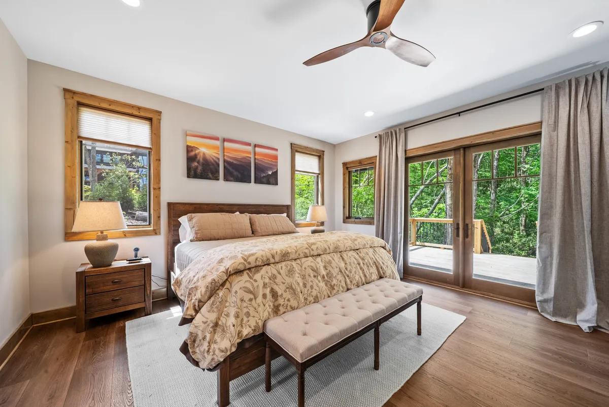 A serene bedroom with a plush king bed dressed in luxury linens sits beside large windows offering picturesque views of nature. Soft lighting from a nightstand lamp and refined wall art create a tranquil and welcoming atmosphere, while the ample natural light underscores the room’s peaceful retreat vibe.