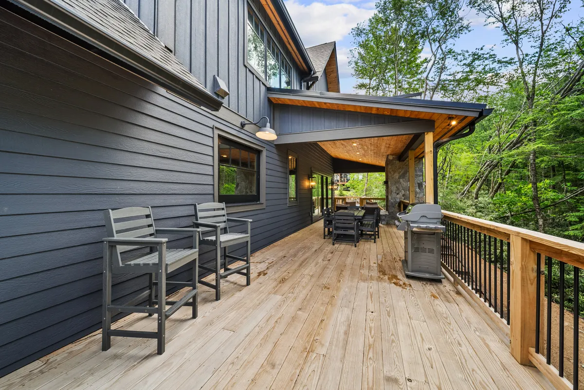 A cozy exterior scene featuring a wooden deck equipped with a barbecue grill and outdoor seating, surrounded by lush greenery and tall trees. The deck offers a tranquil space for families to gather, share meals, and relax after a day of adventure.