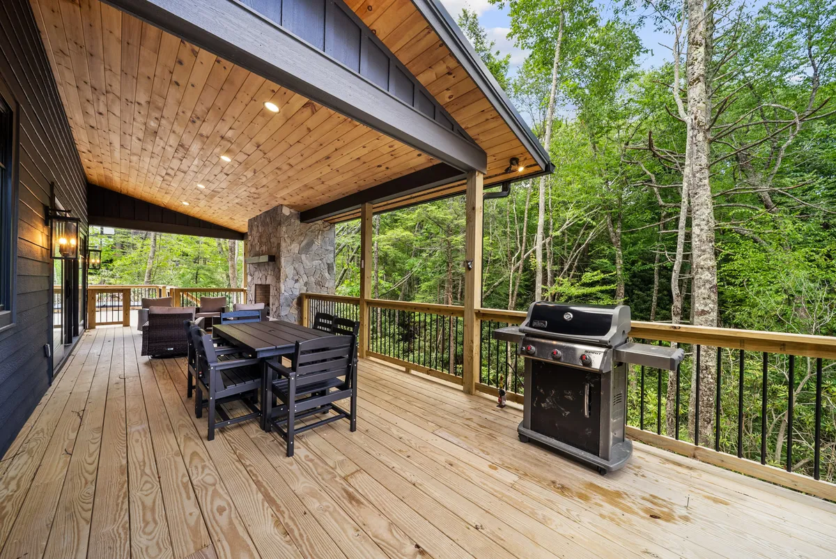 A broad view of the wooden deck, showcasing an outdoor dining table, grill, stone fireplace, and mature trees. The setup is ideal for family gatherings, alfresco dining, and enjoying peaceful mornings in a natural setting.