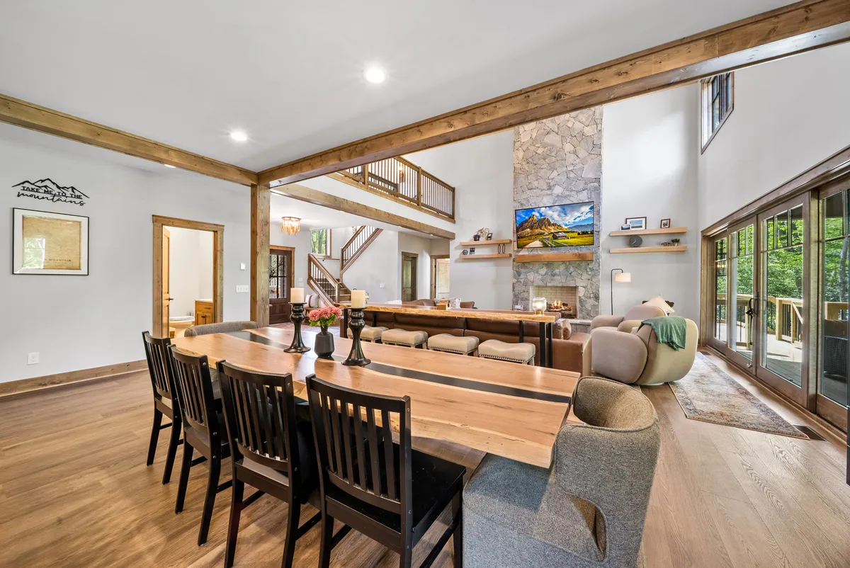 A warm interior scene featuring a dining table and cozy seating area set beside a fireplace, with large windows framing expansive mountain views. The sunlight streams in, hinting at the outdoor scenery and inviting atmosphere for leisurely gatherings.