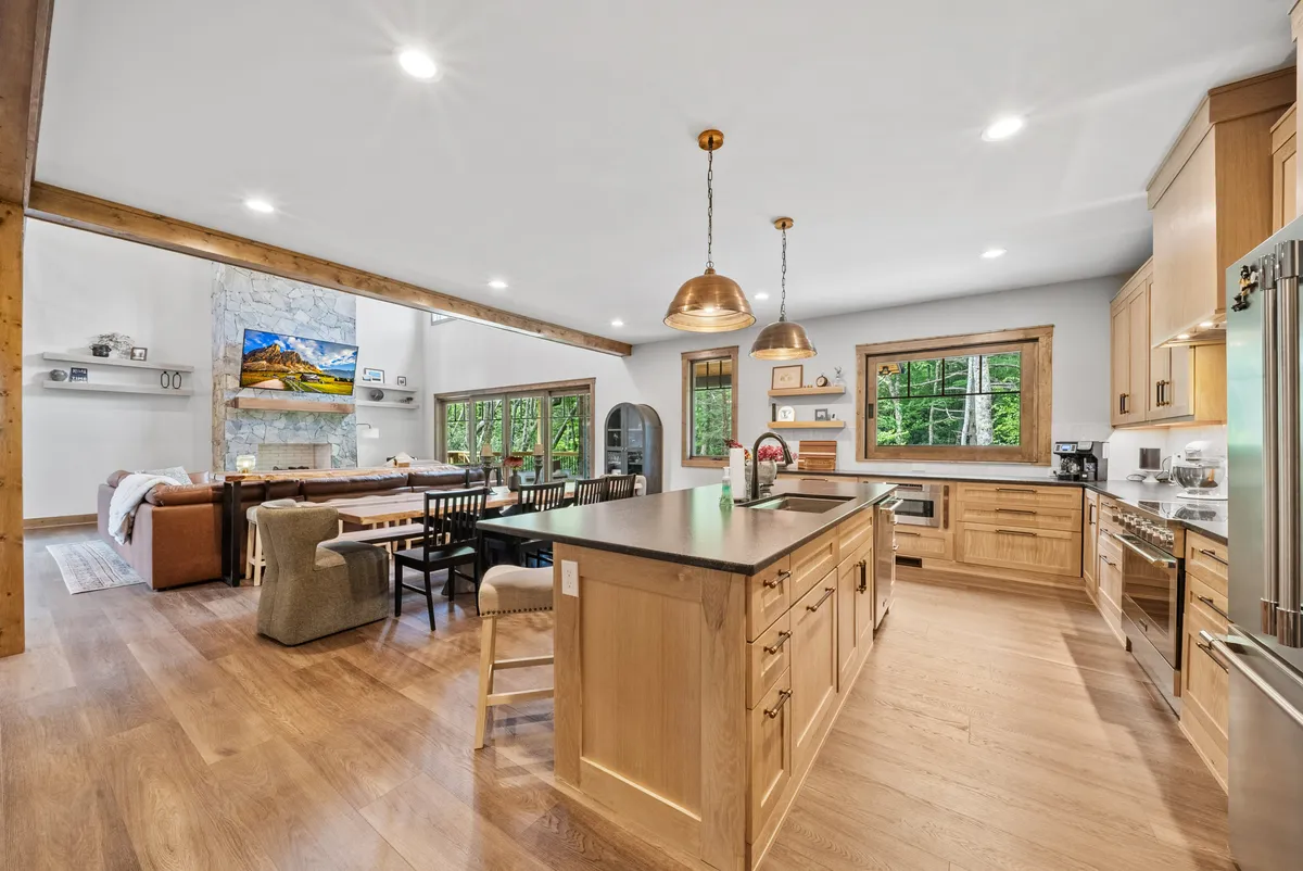 An open-concept kitchen and dining area featuring a prominent kitchen island, a grand dining table, modern fireplace, and expansive windows. The kitchen boasts granite countertops, custom cabinetry, and stainless steel appliances, creating an inviting space for meal preparation and group gatherings.