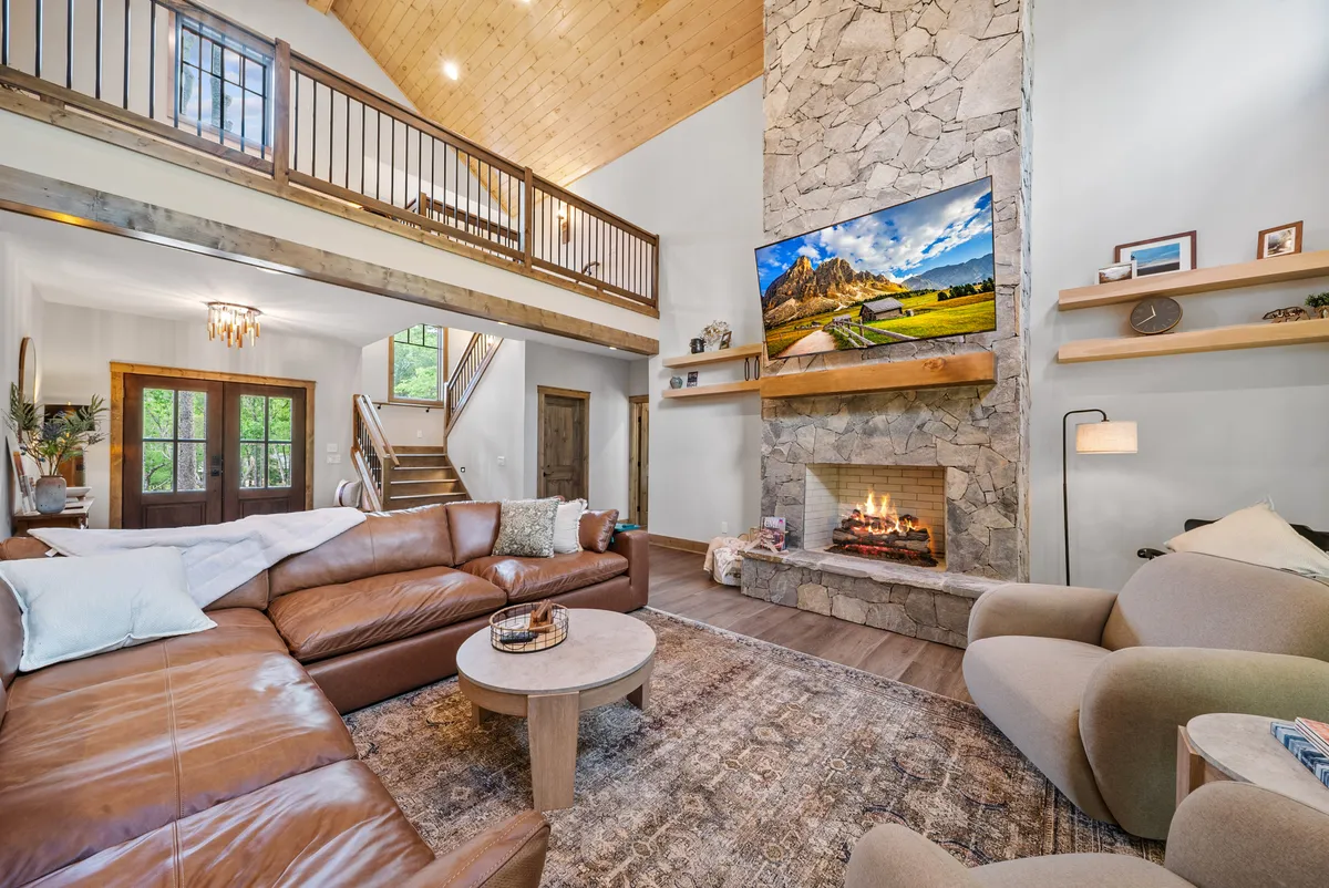 A cozy living area features a plush leather sectional facing a dramatic stone fireplace with a large TV mounted above. Decorative shelves and a wooden staircase add rustic charm, while the space is bathed in warm, welcoming light—inviting guests to relax, enjoy a movie, or share stories by the fire.