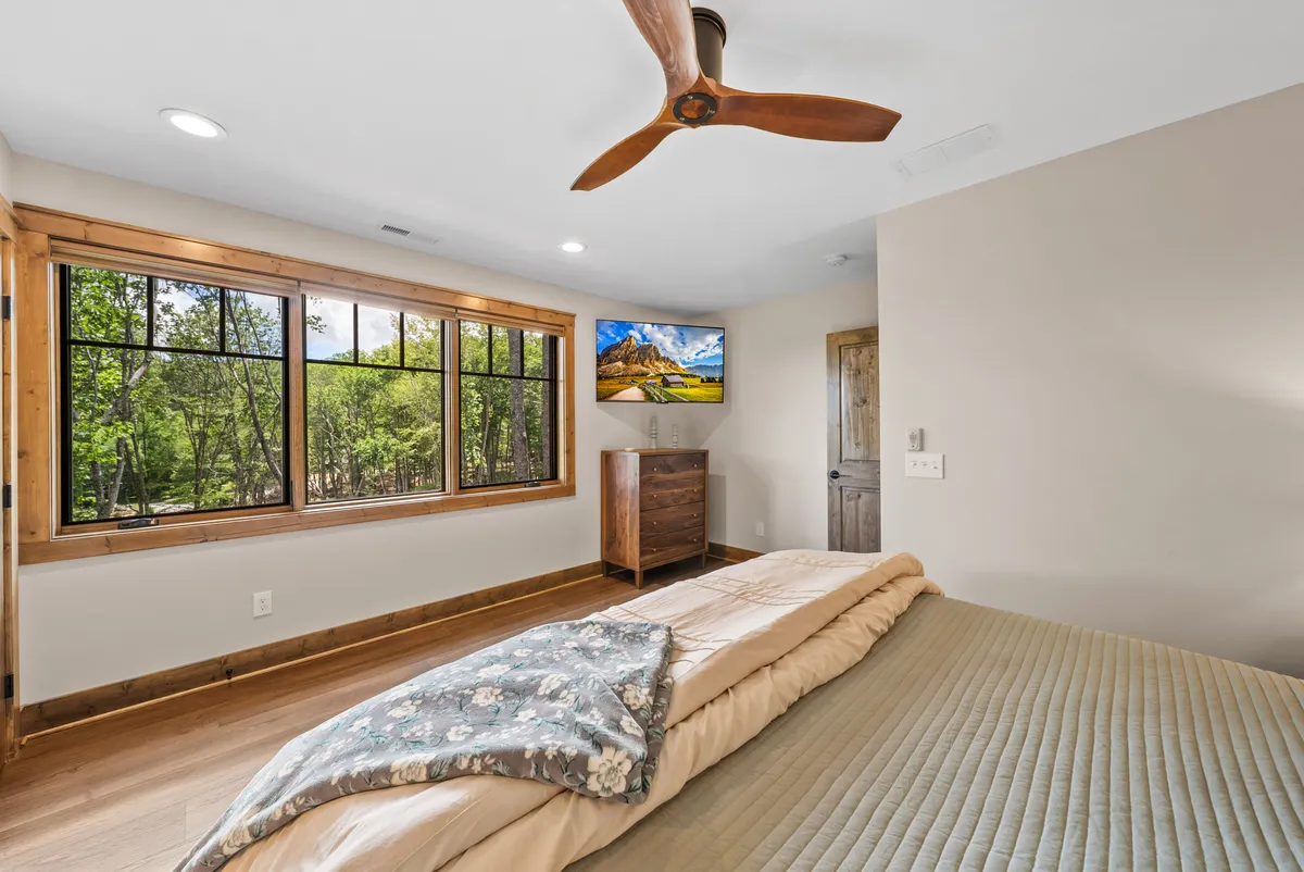 A tranquil bedroom with a comfortable bed dressed in textured bedding, large windows offering views of the outdoors, a ceiling fan for added comfort, and a wall-mounted television. The space feels airy and filled with natural light, inviting guests to relax or enjoy time together.