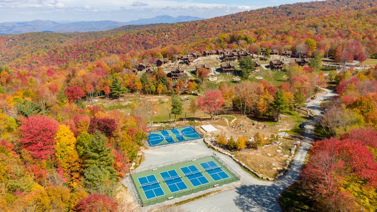 This vibrant exterior image showcases colorful autumn foliage framing well-kept tennis courts, with a dramatic mountain backdrop and nearby residential cabins. The setting hints at active outdoor recreation and a sense of community immersed in nature.