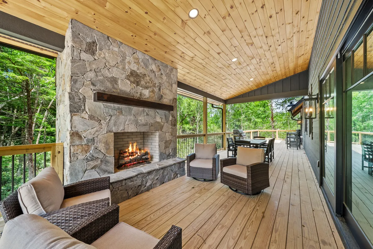 A cozy exterior view of a wooden deck featuring a prominent stone fireplace, comfortable seating area, and lush surrounding greenery. The space is partially covered, inviting relaxation in any weather, and offers a direct connection to nature.