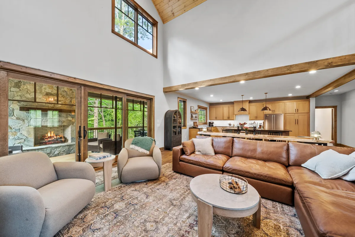 This image showcases a spacious open-concept living area with a large leather sectional sofa centered around a prominent stone fireplace. The vaulted ceilings with rustic wooden beams soar overhead, while expansive windows fill the space with natural light and offer breathtaking views of the Blue Ridge Mountains. The adjacent kitchen area and dining space can be seen flowing seamlessly from the living room, reinforcing a sense of togetherness and effortless entertaining.