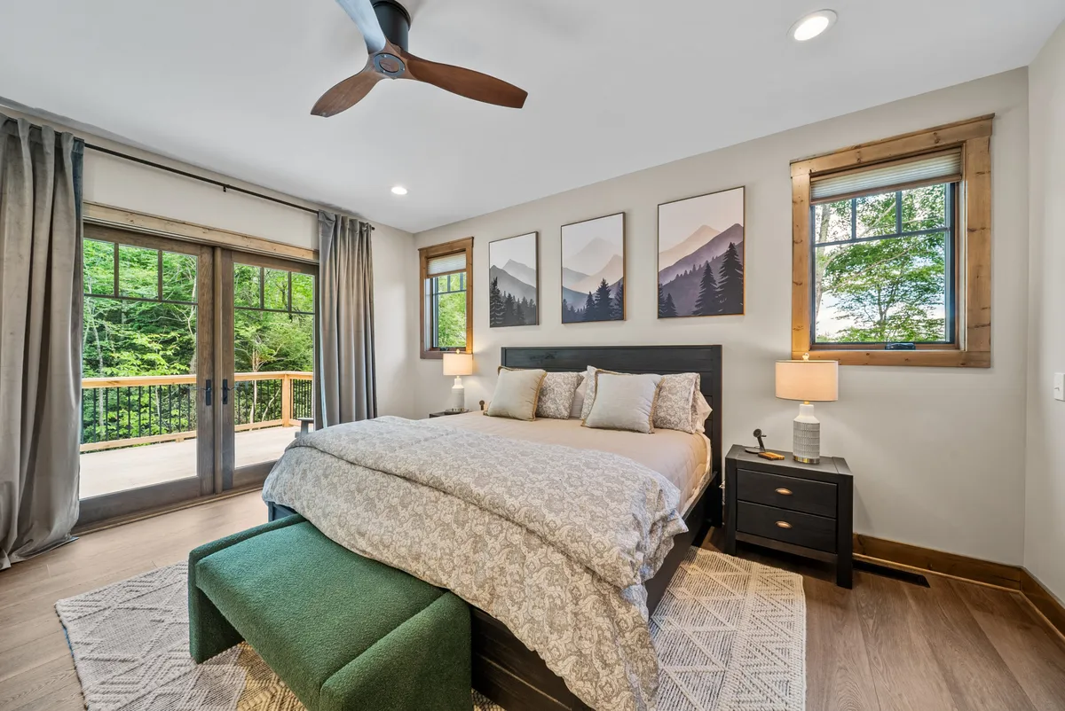 A serene bedroom with a cozy bed, vibrant wall art, and expansive windows framing an outdoor view. The setting is perfect for relaxation, reading, or enjoying a morning coffee with natural light streaming in.