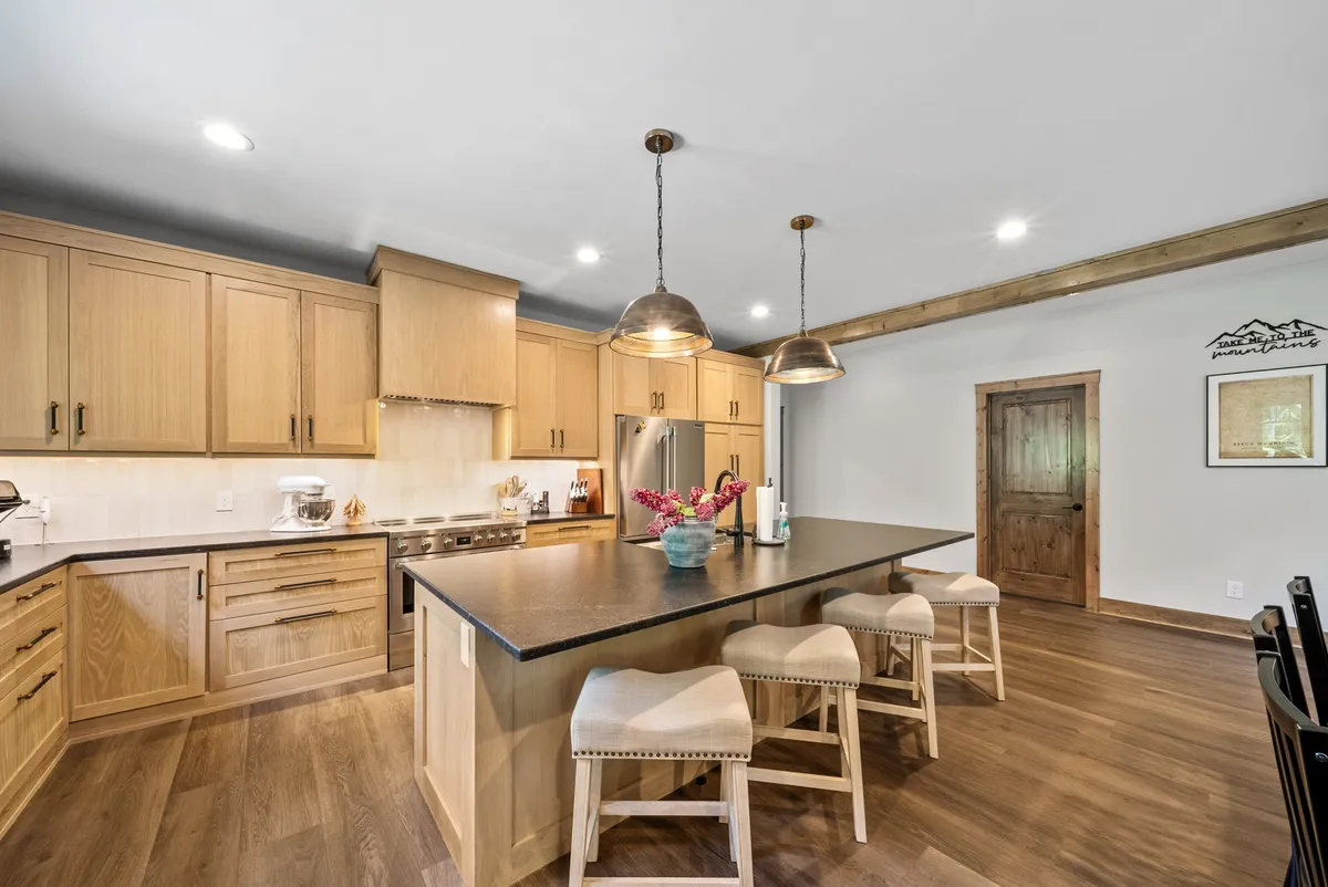 A close-up view of the central kitchen island surrounded by bar stools, accented by hanging light fixtures and rich wooden cabinetry. Stainless steel appliances and ample workspace convey a modern, well-equipped culinary environment ideal for entertaining or casual gatherings.