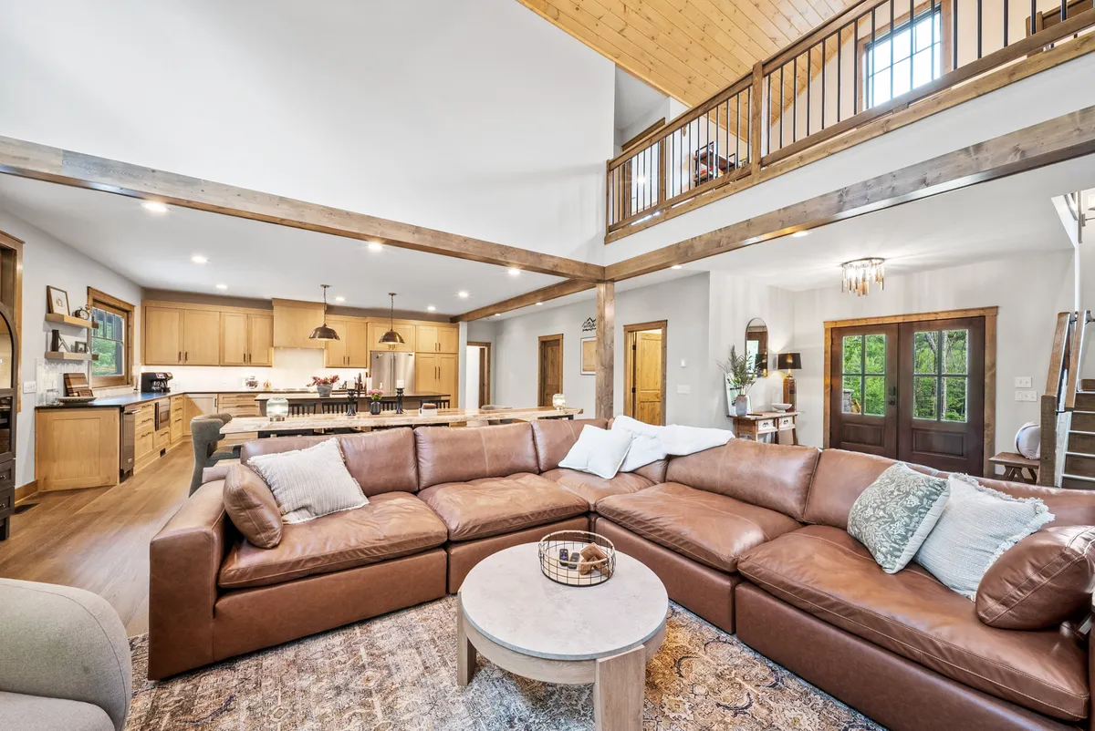 A panoramic view of the central living space reveals a large leather sectional, a wooden dining table, an open kitchen layout, and high ceilings adorned with exposed wooden beams. The arrangement encourages communal gatherings, whether for game nights, shared meals, or simply relaxing together.