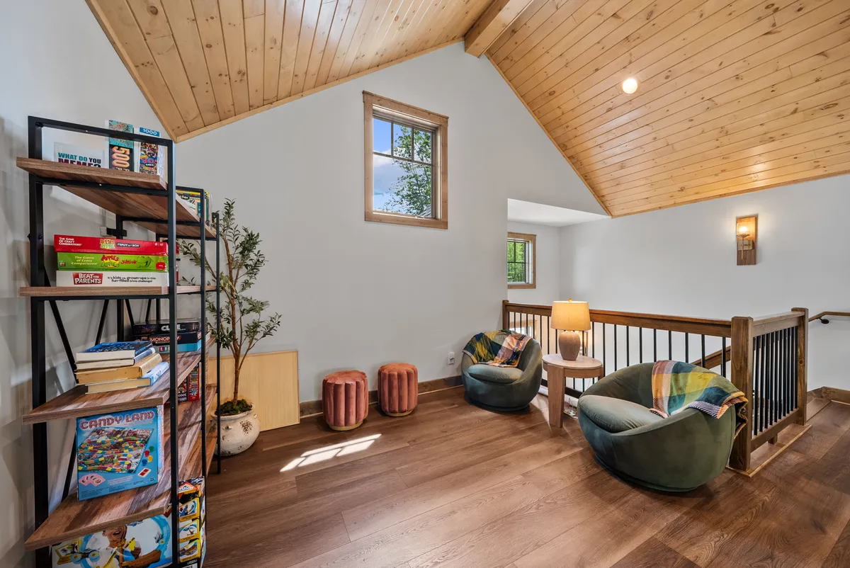 A loft area filled with natural light, featuring a bookshelf lined with games and books, a cozy seating area, and large windows that offer views of the outdoors. The inviting setup makes it ideal for quiet reading or lively family game nights.